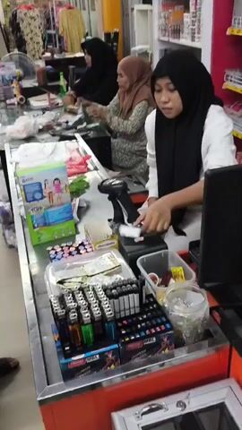 A person wearing a black hijab and a white shirt is working at a checkout counter. They are looking at a computer screen and are in front of a barcode scanner and a cash register. In front of them are various items including lighters, pens, and a box of chocolates. Behind them, two other people wearing hijabs are also working at checkout counters, with one of them holding a green bottle. The environment appears to be a retail store with shelves of products visible in the background.