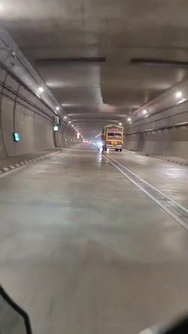 A POV shot from a vehicle traveling through a dimly lit tunnel. The tunnel has concrete walls and ceiling, illuminated by numerous overhead lights. Traffic cones line the right side of the road. A large truck is visible ahead, its headlights shining brightly. The road surface is wet, reflecting the lights. Another vehicle, possibly a motorcycle, is seen at the bottom of the frame, suggesting the perspective is from one.