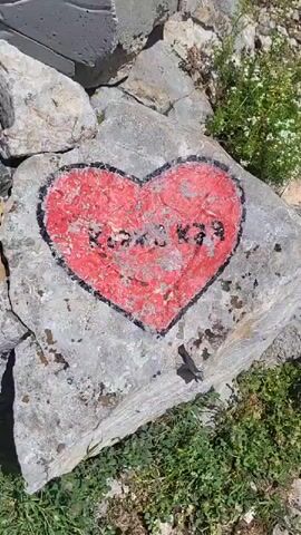 A video pans across a mountain summit marked by a sign reading "врх Коджа кая 597 м." The sign is white and has text and graphics, including a map of the surrounding area with named landmarks and geographical coordinates. A red heart with some writing in a dark color is painted on a large rock in the foreground. There are also several metal poles and a white structure with a graphic depicting a man in athletic wear. The landscape is mountainous with green vegetation and a bright blue sky with white clouds. The video appears to be taken on a sunny day.