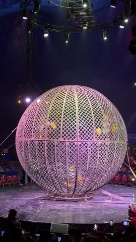 The video shows a large metal sphere on a circus stage. Inside the sphere, several people are riding on motorcycles. The sphere is rotating, and the motorcycles are moving along the inner surface of the sphere. The stage is lit with colorful lights, and an audience is visible in the background.