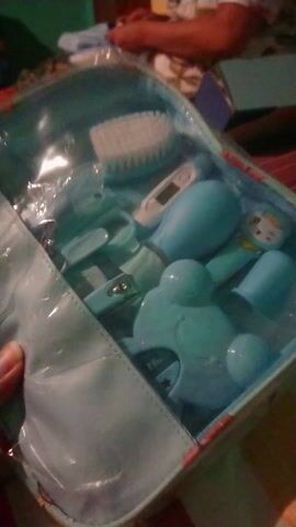 The video opens with a close-up shot of a clear plastic zippered pouch containing a baby grooming kit. Inside, several teal-colored baby care items are visible, including a soft-bristled brush, a digital thermometer, scissors, nail clippers, and a nasal aspirator.

The camera then moves, revealing a light blue mat, possibly a changing mat or playmat, adorned with various cartoon characters such as Mickey Mouse, Winnie the Pooh, and several Tsum Tsum characters. A hand is seen holding this mat. In the blurred background, part of a person, likely a child, wearing a patterned top is briefly visible. The setting appears to be indoors, possibly a home, with soft lighting. The video showcases these baby-related items, suggesting an unboxing or presentation of newborn essentials.