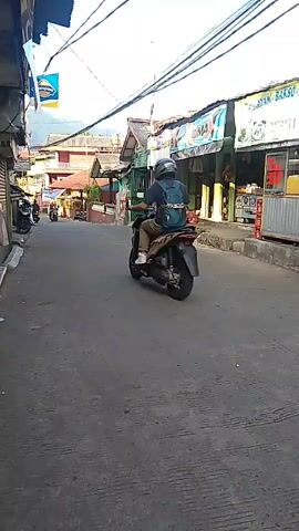 A motorcycle is driving down a street. The street is lined with shops. The shops are open and there are people in them. The motorcycle is a black scooter. The rider is wearing a helmet and a backpack. The street is paved and there are power lines overhead. The sky is blue. There are mountains in the background.