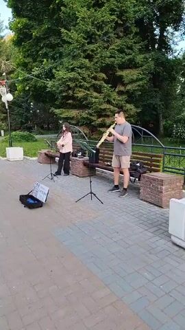 The video shows two people busking in a park in Yoshkar-Ola, Russia. A man is playing a saxophone, and a woman is playing a clarinet. They are standing on a paved walkway in front of a park bench and large trees. There is a music stand in front of each musician, and a small amplifier is placed on the ground. A black open instrument case is on the ground in front of the woman, with sheet music visible inside. The scene appears to be set in the late afternoon or early evening, with soft lighting. The overall atmosphere is relaxed and pleasant, suggesting a casual performance. The weather seems to be good, with green foliage indicating a warm season.
