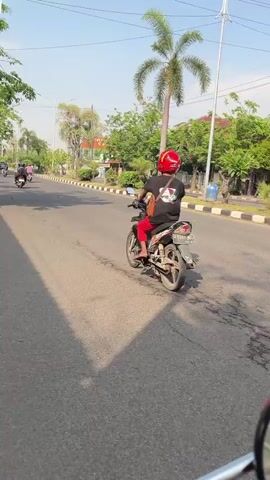 The video shows a person on a motorcycle from behind, wearing a red helmet, a black t-shirt, and red pants. The motorcycle is moving forward on a paved road. The environment is a sunny day with trees and buildings lining the road. Other motorcycles and people can be seen in the background.