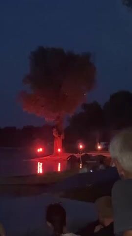This is a video clip of a nighttime scene by a body of water. A large tree is illuminated with red lights, casting reflections on the water. Several red lights are visible around the base of the tree and on the water's surface. In the background, there are silhouetted trees and some distant lights. In the foreground, the backs of several people are visible, as they appear to be watching the illuminated tree. The sky is dark, indicating it is night.