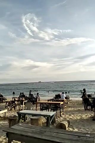 The video shows a crowded beach with people swimming and relaxing on the sand. Several picnic tables and chairs are set up on the beach, some occupied by people. The water is choppy with small waves. In the distance, a small sailboat can be seen. The sky is overcast with some sun breaking through.