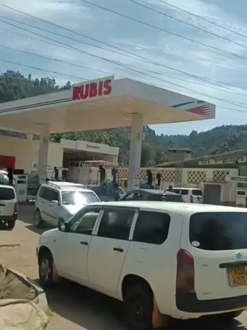 The video shows a Rubis gas station in a rural area. Several cars are parked or queuing at the pumps. The gas station building has a "Service" sign and is located near a green, hilly landscape. Overhead power lines are visible against a partially cloudy sky.  A few people are visible around the vehicles.