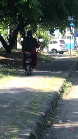 A person riding a motorcycle is seen from behind, traveling down a paved path that runs alongside a grassy area. The person is wearing dark clothing and a head covering, along with maroon pants. The motorcycle has a rear passenger seat and a luggage rack. In the background, there is a white car parked, and further in the distance, a blue and white building is visible. Trees with green foliage line the left side of the path, casting shadows. The path has a curb on the right side, separating it from a road where other vehicles, including another motorcycle, are present. The lighting suggests it is daytime, with sunlight filtering through the leaves.