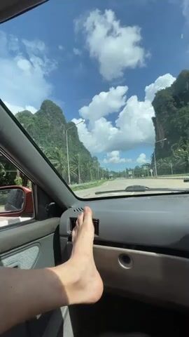 The video begins with a close-up of a person's bare foot resting on the dashboard of a car. The car appears to be in motion, as indicated by the blurred scenery outside the windshield. The dashboard is gray and shows a vent and a compartment. The windshield displays a bright blue sky with fluffy white clouds and lush green mountains in the distance, lined with palm trees. A road is visible in front of the car, with other vehicles and streetlights. A smartphone mounted on a holder displays a navigation map. The perspective then shifts to show the view from the side window, revealing the road, palm trees, and mountains, with a red side mirror in the foreground. The video appears to be shot from inside a car driving on a road with a scenic mountainous landscape.