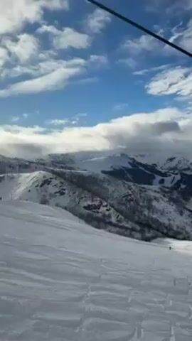 The video shows a snowy mountain landscape with a clear blue sky and scattered clouds. A ski slope is visible, with tracks in the snow. There are other mountains in the background, some with trees and some covered in snow. A ski lift cable runs overhead. A few people are skiing down the slope.