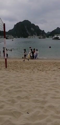The video captures a beach scene with a group of people.  A sandy beach is in the foreground, marked by a red and white volleyball net pole. In the middle ground, several individuals stand and interact near the water's edge. They wear varied clothing, including white and red robes. One person in athletic wear jogs toward the left. In the background, a body of water is dotted with cruise ships.  The horizon is lined with mountainous land. The sky is overcast.