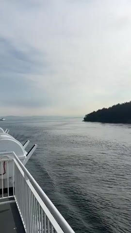 The video shows a view from a ferry as it travels through calm waters. The ferry has a white railing in the foreground. On the right side of the screen, a densely forested island with a hilly coastline is visible. The sky is partly cloudy with the sun shining brightly at times, casting reflections on the water. The ferry is creating a wake behind it. The overall scene is serene and depicts a journey through a natural, coastal environment.