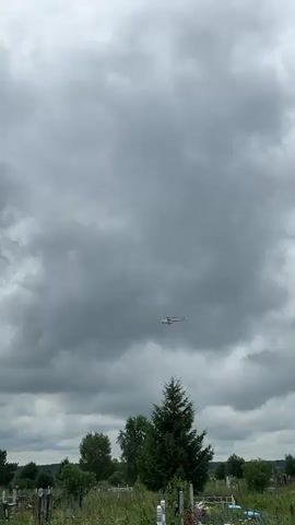 A helicopter flies across a cloudy sky over a grassy area with trees.