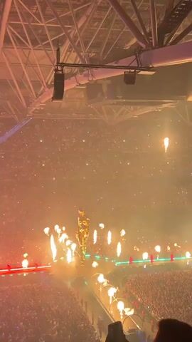 A large indoor concert venue is filled with an audience. The stage features a golden statue-like figure surrounded by pyrotechnics. The audience is visible in the foreground and fills the seats. Lights, including green and blue lasers, illuminate the stage and crowd. The ceiling structure of the venue is also visible, with speakers mounted overhead.