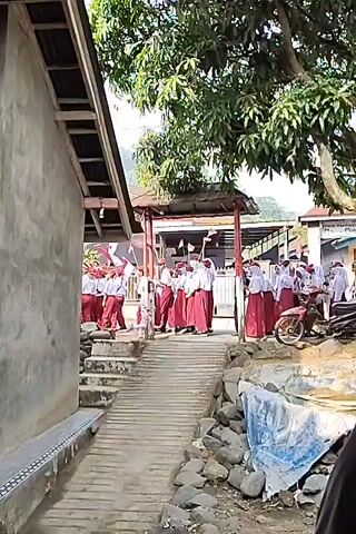 A group of students in red and white uniforms are gathered on a ramp leading to a covered area. A few adults are present among them. A motorcycle is parked to the right of the ramp. The environment appears to be outdoors, with trees and buildings visible in the background. The scene is brightly lit, suggesting daytime.