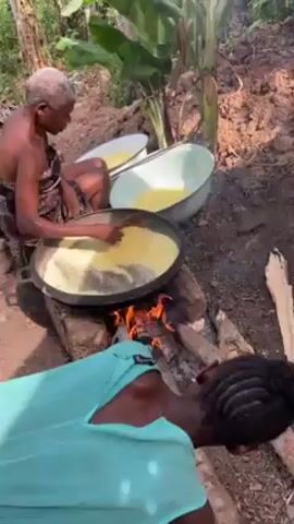 Two women are preparing food outdoors. One woman kneels beside a fire, stirring something in a large pan. The fire is built directly on the ground, and she adds pieces of wood to it. The second woman sits behind the first, also stirring something in a large pan. There are two other pans or basins nearby, both containing a yellow liquid. The surroundings are dirt, wood debris, and tropical foliage.