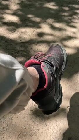 The video shows a person's foot clad in a hiking shoe and black sock with pink trim, while the person sits outdoors on a sandy surface dappled with tree shadows. They are wearing gray pants.  A black dog walks in the background.