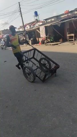 A man is walking down a paved street, pulling an empty, metal-framed cart. The cart has four wheels and two handles. The man is wearing a red and yellow tank top, black pants, a red baseball cap, and black sandals. He is pulling the cart away from the camera. The street is lined with shops and other businesses. There are people, motorcycles, and cars visible in the background. Overhead are power lines.