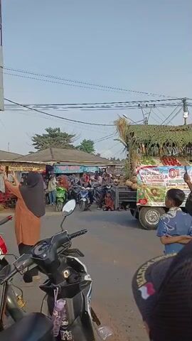 A street scene with many people and motorcycles. A truck is parked on the right side of the street, loaded with produce. A woman in an orange headscarf and hijab is standing on the left side of the street, holding up her phone, possibly taking a picture or video. Several other people are gathered around the truck, some appearing to be interacting with the produce or the people on the truck. The environment appears to be outdoors, with a clear sky and some trees visible in the background. The overall impression is that of a bustling market or a community event.