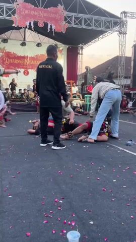 The video shows a group of people on a stage. The foreground is covered in fallen rose petals. A person wearing a black shirt with "OFFICIAL" in yellow lettering on the back stands facing away from the camera, observing the scene. In front of them, several individuals are lying on the ground, some appearing to be in a theatrical performance. Two people are actively assisting individuals who are on the ground, one person is in blue jeans and a light-colored shirt, kneeling and reaching towards someone on the ground, and another person in a green shirt is also leaning over. There are other performers and people on the stage in the background. A banner with text is visible on the stage backdrop. The lighting suggests it is late afternoon or early evening.