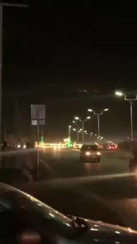 The video shows a night-time view from a vehicle moving along a road. Streetlights illuminate the road, and other vehicles are visible in the distance. The road appears to be divided by a median. There is a signpost on the side of the road. It is dark outside, and the only light comes from streetlights and the headlights of vehicles. The camera is positioned low to the ground, possibly inside a car.