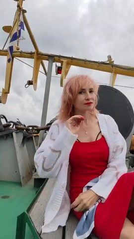 A woman with short, light-pink hair and red lipstick is sitting on the deck of a boat. She is wearing a red dress and a white cardigan with a heart pattern. The woman is looking towards the camera, her head tilted slightly and one hand touching her neck. Her hair is blowing in the wind. In the background, there is a metal structure, chains, and a flag. The sky is overcast. The scene appears to be outdoors, likely on a body of water, given the presence of the boat and the flag. The woman's expression is neutral or slightly pensive.