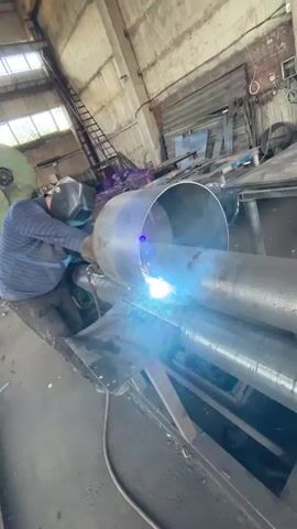 The video shows a person wearing a welding mask and protective clothing welding a large metal cylinder. The cylinder is resting on a rolling machine with multiple rollers. Sparks and bright light are emanating from the welding torch. The environment appears to be a workshop with industrial equipment visible in the background, including a ladder and metal sheeting. The welding process is ongoing.