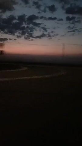 The video shows a view from a moving vehicle, likely a car, looking out at a dusk or dawn sky. The sky has a gradient of colors, with pinks and oranges near the horizon and darker blues and grays higher up, dotted with scattered clouds. The ground in the foreground is dark and appears to be a road or field. There are faint lines visible on the road, suggesting lane markings or tire tracks. In the distance, there are power lines and possibly some hazy structures. The overall impression is a fleeting glimpse of a vast, open landscape during twilight.