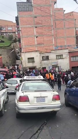 The video shows a street scene with a large group of people gathered near parked cars. The cars include taxis, indicated by roof signs. A brick building is visible in the background, and a hillside with greenery rises on the left. The people are standing close together, some wearing face masks. The sky is overcast. There's general activity, suggesting a crowded public area.