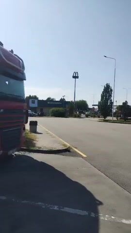 The video shows a wide shot of a truck stop or rest area under a clear blue sky. In the foreground, the side of a red semi-truck is visible on the left. The road surface is asphalt, with a yellow painted line on the left edge. In the middle ground, a McDonald's sign is visible, along with a building that appears to be the restaurant. Several semi-trucks are parked in the area. A blue semi-trailer truck is visible on the right side of the road, and another grey trailer is behind it. Further in the distance, more trucks are parked. Tall streetlights are present throughout the area. The overall impression is a typical highway service area.