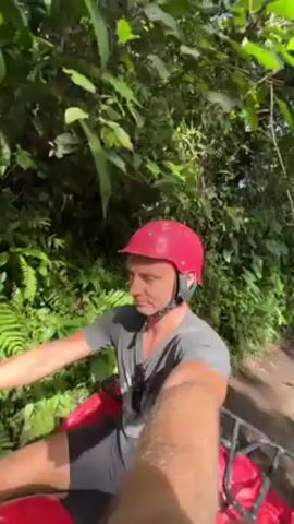 A man is seen riding a red ATV through a lush green forest. He is wearing a gray t-shirt, black shorts, and a pink helmet. He is smiling and looking directly at the camera, which appears to be mounted on the ATV, capturing a first-person perspective. The forest is dense with thick foliage, including ferns and various trees, with sunlight dappling through the leaves. The ATV is moving along a dirt path.