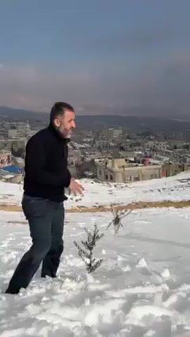 A man is throwing snowballs in a snowy landscape. He is wearing a black sweater and blue jeans. He is holding two snowballs in his hands and throwing them in the air.  Behind him is a snow-covered field with a small tree. In the background, there is a town with buildings and snowy hills. The sky is blue with some clouds.