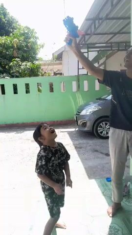 A child is standing in front of a green wall, looking up at an adult who is holding a blue toy gun and aiming it upwards. The adult is wearing a dark t-shirt and light-colored pants. The child is wearing a dark, patterned outfit. Behind them, a car is parked. The scene appears to be outdoors, with sunlight casting shadows. The adult lowers the toy gun and then crouches down.