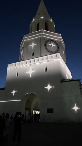 The video shows a tall, white building at night, illuminated with white lights. The building has a pointed tower with a clock face on one side. White star-shaped lights are projected onto the building's facade. People are walking in front of the building, passing through an arched entrance. The sky is a dark blue. The camera angle is low, looking up at the building.
