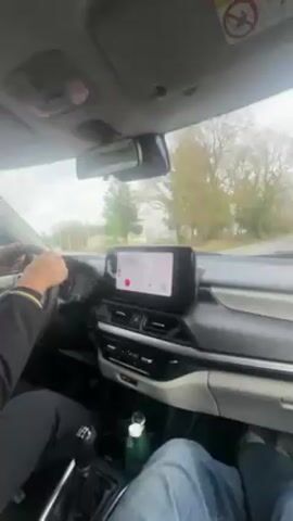 The video shows the interior of a car driving down a road. The driver, whose hands are visible on the steering wheel, is wearing a dark-colored jacket with a gold stripe. The passenger is wearing blue jeans. A bottle of water sits in the center console. The car's dashboard features a large infotainment screen. The view through the windshield shows a road flanked by trees, suggesting a rural or suburban setting. The sky is overcast, indicating a cloudy day.