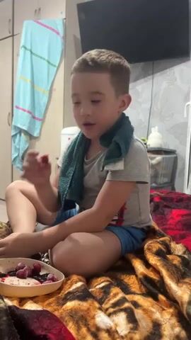 A young boy is sitting cross-legged on a patterned blanket, eating berries from a white dish. He is wearing a grey t-shirt, blue shorts, and socks. A green towel is draped around his neck. He takes a berry from the dish and puts it in his mouth, then chews and swallows. He then reaches for another berry, picks it up, and brings it to his mouth. He then puts the towel around his neck. In the background, there is a closet with a light blue towel hanging on it, a television, and some shelves with items on them.