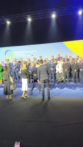 This media file captures a moment during the Ukraine Recovery Conference held in Gdańsk, Poland. The scene is set on a large stage in a conference hall, indicated by the professional lighting rig overhead and the expansive blue backdrop. Prominently displayed on the backdrop is the conference logo, which reads "UkraineRecovery Conference" followed by "23-24 June 2023, Gdańsk, Poland." A large group of people, predominantly men in suits and some women in formal attire, are gathered on tiered seating behind a blue carpeted area. They appear to be dignitaries, politicians, or delegates attending the event.

In the foreground, a few individuals are walking on the stage. Two women in dark and white attire are positioned on the left, seemingly interacting with each other or preparing to join the group on the tiered seating. A man in a dark suit is walking away from the camera towards the larger group, his posture suggesting purpose. Another person's hand holding a smartphone is visible at the bottom of the frame, indicating that the media was likely captured by an attendee.

The atmosphere appears formal and official, befitting a high-level international conference focused on Ukraine's recovery. The lighting is bright and professional, suggesting the event is taking place indoors during daylight hours. There are no overt displays of emotion, but the overall impression is one of serious engagement and international cooperation. The presence of national flags behind the main text on the backdrop further underscores the international nature of the gathering.
