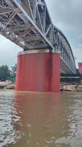 The video shows a view from a body of water looking up at a large metal bridge. The bridge has a complex steel structure, and one of its massive support pillars is painted a vibrant red. The water appears murky and brown, suggesting it may be a river. In the background, on the far bank, there are some trees and buildings, and several small boats are docked. The sky is overcast with some visible clouds. The camera perspective is low, emphasizing the scale of the bridge. There are no people visible in the video, and the primary action is the camera's slow upward and sideways movement, capturing different angles of the bridge. The overall impression is one of industrial infrastructure situated within a natural environment.