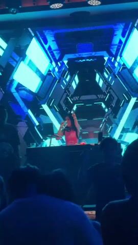 A woman in a pink top and headphones stands behind a DJ booth, holding a drink and speaking into a microphone. She is in a dimly lit room with a large screen behind her displaying a geometric, blue and black pattern. Several people are visible in the foreground, silhouetted against the light. The woman is gesturing with her hands as she speaks.