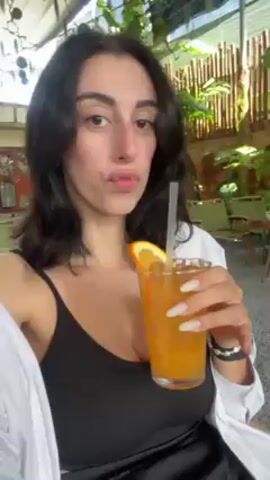 The video shows a dark-haired woman holding a glass of orange liquid with a straw and orange slice. She is sitting in what appears to be a partially outdoor restaurant with hanging plants and bamboo decor visible in the background. She's wearing a black tank top with a white button-down shirt. She takes a sip of the drink through the straw.