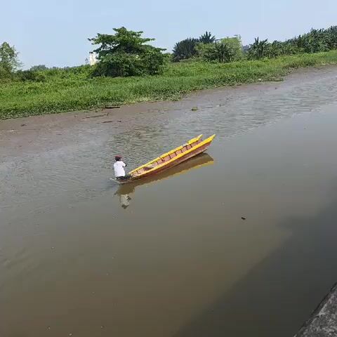 A person in a white shirt and dark pants stands in the water, pushing a yellow and red boat. The boat is in a muddy river with green foliage and trees on the bank. The sky is clear and blue.