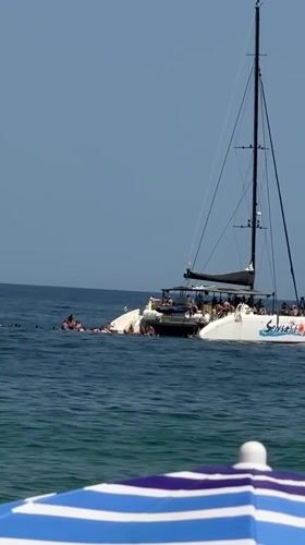Packed summer beach day in Badalona, catamaran anchored offshore