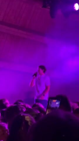 The video shows a concert with a performer on stage under purple lighting. The performer is singing into a microphone and gesturing with their hands. A crowd of people is visible in the foreground, with some holding up their phones to record the performance. The concert appears to be taking place outdoors or in a large venue with exposed wooden beams on the ceiling. The overall atmosphere is energetic and celebratory.