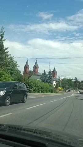 This is a first-person perspective video taken from inside a car, likely a dashboard camera, as it drives through Ostrów Mazowiecka, Poland. The camera is angled slightly upwards, capturing the view through the windshield and side windows.

The scene is an urban or semi-urban environment on a bright, sunny day with scattered clouds. The dominant structures visible are a large, red-brick church with multiple spires and a green roof, surrounded by trees and other buildings. A paved road with white lane markings stretches ahead, indicating the car is in motion. There are also power lines visible overhead. Along the right side of the road, there's a sidewalk, streetlights, and more vegetation, including tall evergreen trees. Further down the road, other vehicles and buildings can be seen. The video captures the car passing by the church and continuing its journey.

There are no people clearly identifiable as subjects of the video, but the presence of vehicles implies human activity. The overall mood appears to be that of a casual drive through a town on a pleasant day. There is no visible text in the video.