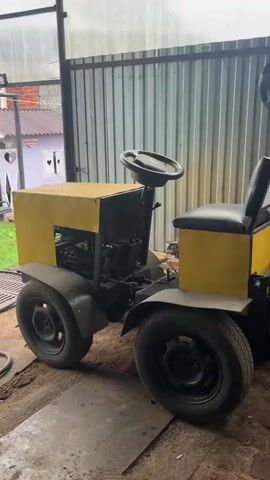 The video displays a yellow and black mini-tractor. The tractor is stationary inside a workshop, with a corrugated metal wall behind it. The camera pans to the right, revealing a blue tractor with a cabin. This tractor is also stationary and appears to have some sort of loader attachment on the front. The workshop appears to be cluttered with various tools and equipment, including what looks like a welding machine. The lighting is dim and appears to be natural light coming from above, possibly through a transparent roof.