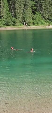 This media file captures a serene natural scene at Lago di Braies (Pragser Wildsee) in Italy. The primary subjects are two individuals swimming in the crystal-clear, emerald-green water of the lake. One person is closer to the foreground, with their head and shoulders visible above the water. The second person is further out in the lake, appearing to be swimming or floating.

The setting is a breathtaking mountain lake, characteristic of the Dolomites region. Majestic, rugged mountains rise steeply behind the lake, covered in dense green pine forests. The shoreline is a mix of pebbles and sand, with some scattered rocks. Several other people are visible on the shore in the background, enjoying the scenery or perhaps preparing for activities. A few small boats are also present on the water in the distance.

The time of day appears to be daytime, likely late morning or early afternoon, given the bright sunlight casting shadows and illuminating the scene. The weather seems pleasant and sunny, contributing to the inviting atmosphere for swimming. The overall mood is one of tranquility, natural beauty, and recreational enjoyment. There is no visible text in the media file.