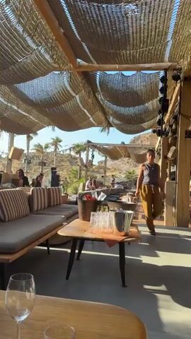 A woman wearing a gray tank top and tan pants walks past a table in an outdoor seating area. The table has a wooden top and black legs, and it holds a bucket of ice with bottles in it, as well as glasses and napkins. In the background, there are several people sitting on couches and tables. Palm trees and hilly terrain are visible beyond the seating area. The area is covered by a woven canopy.