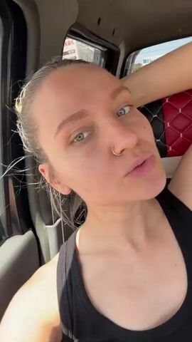 A woman is in the passenger seat of a car, looking up at the camera. She is wearing a black tank top and has a nose ring. Her hair is pulled back. She is making a kissy face. The car seat behind her has a black and red quilted pattern. Outside the car window, a building with the word "LUXOIL" on it is visible. The woman raises her hand to the back of her head.