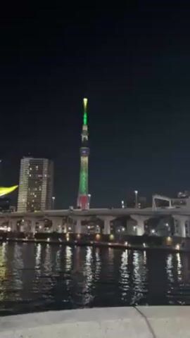 The video shows the Tokyo Skytree tower at night. The tower is lit up with green, yellow, and red lights. There is water in the foreground and buildings in the background. There is a bridge in the midground.