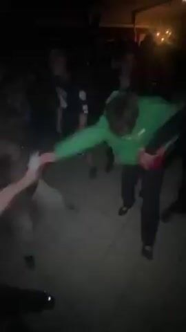 The video shows a group of people moshing or slam dancing at an outdoor event. The people are a mix of genders and are dressed in casual clothing. The light is dim, but there is a building visible in the background with string lights. The floor is tiled.